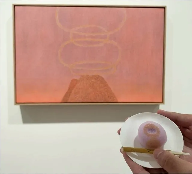 ピンク色の抽象絵画の前に、その色彩と形態を忠実に再現した練り切りの和菓子を小皿に載せて掲げている様子