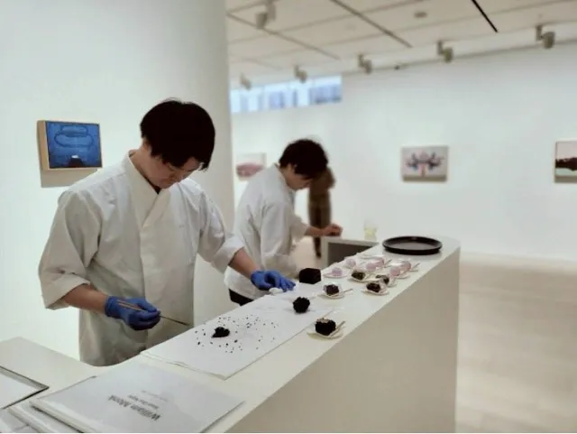 ギャラリー内のカウンターで白衣の和菓子職人たちが練り切りを仕上げている制作風景。壁面には複数の絵画作品が掛かっている