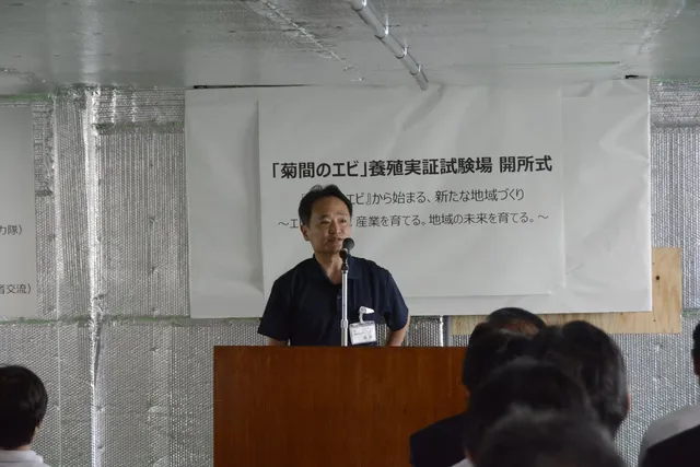 「菊間のエビ」養殖実証試験場の開所式で挨拶する関係者。背後の横断幕に『「菊間のエビ」から始まる、新たな地域づくり ～エビを育てる。産業を育てる。地域の未来を育てる。～』と書かれている