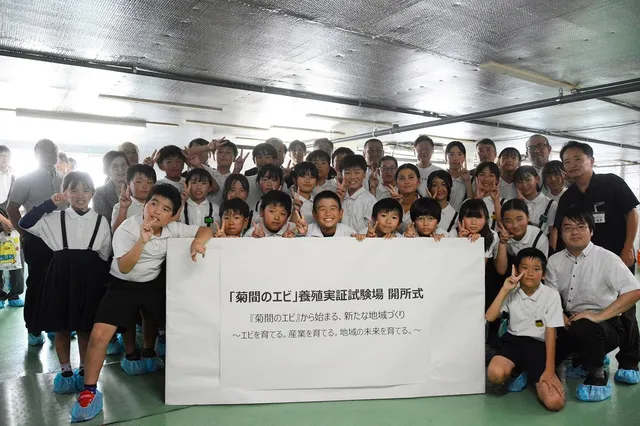 開所式に参加した地元の小学生たちと関係者の集合写真。子どもたちが笑顔でピースサインをし、横断幕『「菊間のエビ」養殖実証試験場 開所式 ～エビを育てる。産業を育てる。地域の未来を育てる。～』を囲んでいる