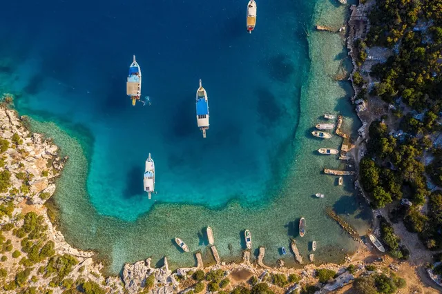 トルコ沿岸の入り江を真上から捉えた空撮写真。エメラルドグリーンの浅瀬に浮かぶ木造ヨットと、岩場に係留された小舟が見える