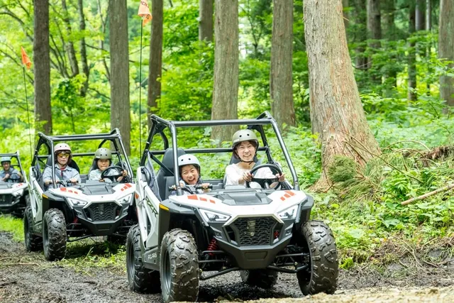 白馬五竜の森の中をPolaris RZRバギーで走る親子連れ。先頭車両では母親がハンドルを握り子どもが助手席に座る。背後の杉林の緑が深い。