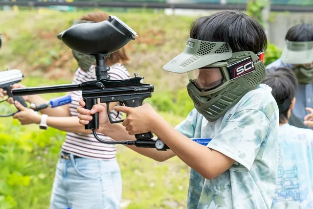 防護マスクを着けた子どもがペイントボール用マーカーを構えている。背後にはもう一人のプレイヤーも見える。