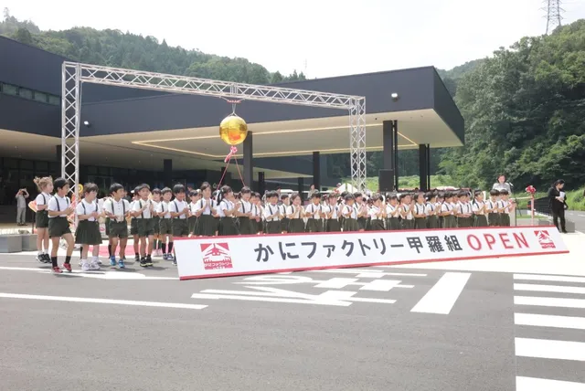 「かにファクトリー甲羅組 OPEN」の横断幕の前に整列する地元の園児たちと、背後に広がる施設と緑の山並み