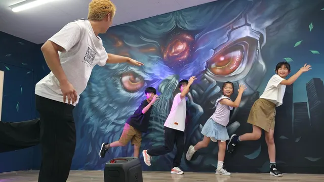 裏トシマのダンススタジオで、金髪の男性インストラクターが子どもたち4人にダンスを教えている。背景には巨大なモンスターの壁画