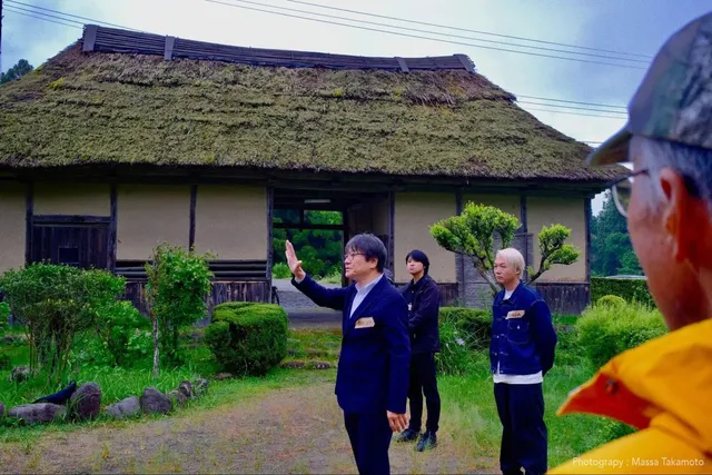 北條家住宅の茅葺き屋根の主屋を背景に、プロジェクト関係者が蔵の修復方針について説明している様子。Photography: Massa Takamoto