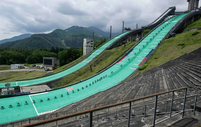 白馬ジャンプ競技場のラージヒルとノーマルヒルの全景。緑色の人工芝が敷かれた夏仕様の斜面を、西窪友海さんが前輪一本で駆け下りる連続写真が合成で示されている