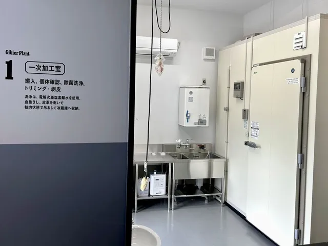 MUBGジビエ解体加工施設の一次加工室。搬入・除菌洗浄・トリミング・剥皮の工程説明が壁に掲示され、ステンレスの作業台と冷蔵庫が並ぶ