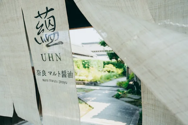 蘊（UHN）奈良マルト醤油の暖簾が風にたなびく入口。奥に緑豊かな庭と古民家の建物が見える