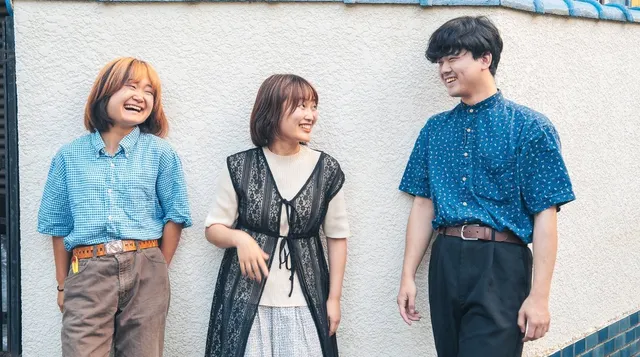 白い壁の前に並んで笑い合う友人3人の全身写真。左からギンガムチェックのシャツの女性、黒いレースのベストを着た女性、青い柄シャツの男性