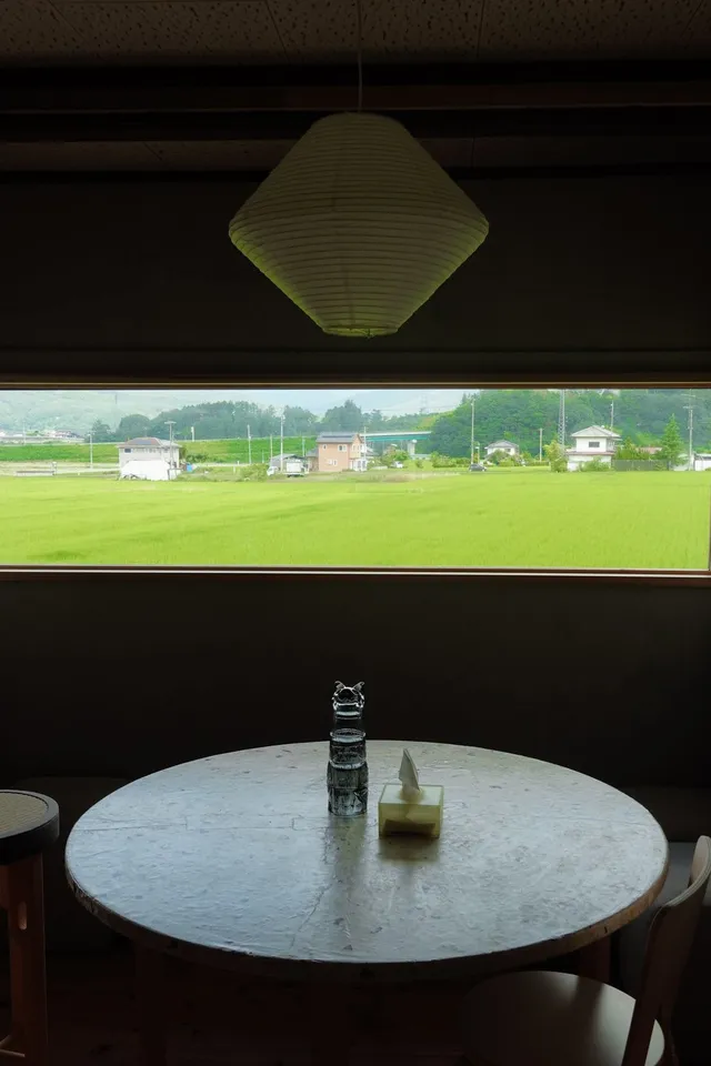 暗い室内から横長の窓越しに広がる青々とした田んぼの風景。丸テーブルの上にはガラスの置物と小さなティッシュケースだけが置かれている