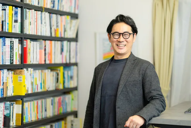 本棚の前に立ち、笑顔でこちらを見るNIJINアカデミー校長・星野達郎さん