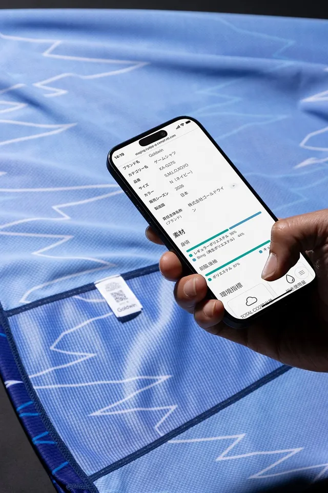 ユニフォームの生地に触れながらスマホでデジタルプロダクトパスポートの情報を読み取る手元。素材構成やCO2排出量の画面が表示されている