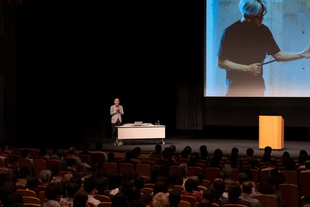 壇上でスクリーンに坂本龍一の映像を映しながら講演する登壇者と、耳を傾ける満席の観客