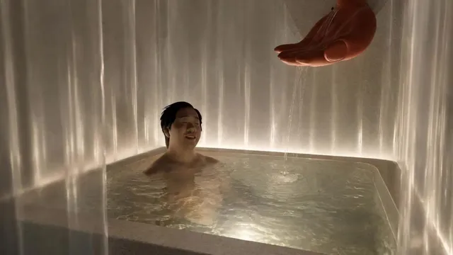 浴槽に浸かり目を閉じてリラックスする男性の上から手のオブジェが湯を注いでいる演出