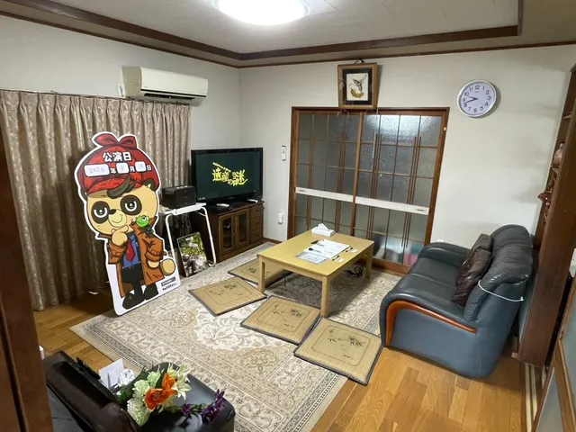 謎解きゲームの会場となる空き家の居間。座布団や机が並び、たぬき探偵ジェリーのパネルやテレビが置かれた生活感の残る空間