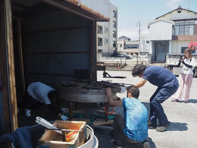屋外で数人がかりで錆びついた大きな回転式せんべい焼き機械を運び出す様子
