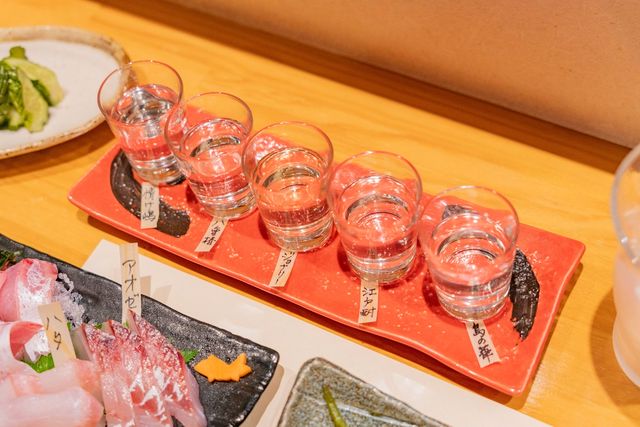 赤い器に並んだ島焼酎の5種利き酒セットと島魚の刺身。ラベルにはそれぞれの銘柄名が記されている